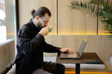 Gözlüklü ve dizüstü bilgisayarlı şık sakallı bir adam kahve dükkanında bir masada oturuyor ve uzaktan çalışıyor. 