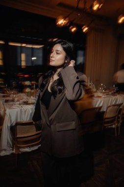 photo of a stylish brunette in an oversized pantsuit with a jacket against the background of a restaurant interior with blurred lights posing stylishly for a photo clipart