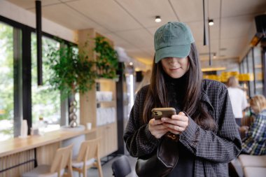 Elinde telefonuyla gülümseyen çekici bir kız bir kahve dükkanının önünde durmuş arkadaşlarını ya da siparişini bekliyor ve telefonu kullanıyor. 