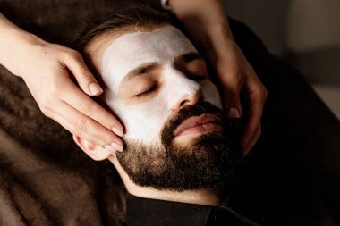 Yakışıklı sakallı adam yüz maskesi takıyor ve yüz masajı yapıyor, yakın plan. Güzellik prosedürü