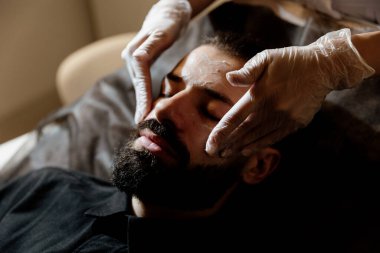 Yakışıklı sakallı adam yüz maskesi takıyor ve yüz masajı yapıyor, yakın plan. Güzellik prosedürü