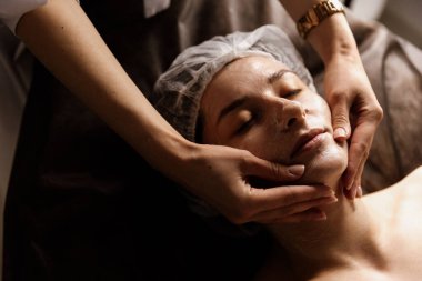 Güzel bir kadının yakın plan portresi, yan ışığı olan bir güzellik merkezinde nemlendirici ile yüz masajı yaptırıyor. Cilt bakımı için doğal kozmetik ürünleri. Sağlık hizmetleri, güzellik