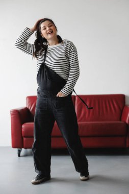 Hamile bir kadın yerde oturuyor, kırmızı bir kanepeye yaslanıyor, çizgili bir gömlek ve siyah pantolon giyiyor, karnına bakıyor..