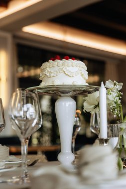 Üzerinde ahududu olan zarif beyaz pasta, uzun bir stantta sergileniyor, şarap kadehleri ve mum ile çevrili, sofistike bir yemek ortamında hazırlanıyor..