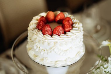 Üzerine taze çilek konmuş güzel dekore edilmiş beyaz bir pasta, cam pasta standına konmuş. Pasta karmaşık buz kalıpları içeriyor ve bulanık bir arkaplan üzerine kurulmuş..