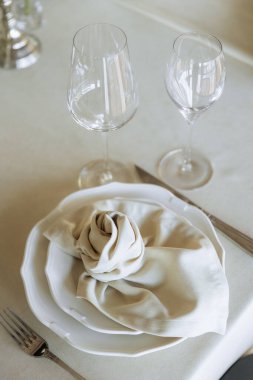 Beyaz tabaklı zarif bir masa, gül gibi katlanmış bir peçete, ve krem masa örtüsü üzerinde iki şarap bardağı..