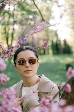 Bahar ceketi, pantolon ve güneş gözlüğü giymiş bir kadın, mülkünün parkında yürürken fotoğraf için poz veriyor.
