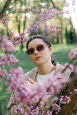 Bahar ceketi, pantolon ve güneş gözlüğü giymiş bir kadın, mülkünün parkında yürürken fotoğraf için poz veriyor.