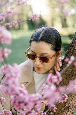 Bahar ceketi, pantolon ve güneş gözlüğü giymiş bir kadın, parkta çiçek açan bir kiraz ağacının arka planında zarif bir şekilde poz veriyor. 