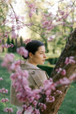 Bahar ceketi, pantolon ve güneş gözlüğü giymiş bir kadın, parkta çiçek açan bir kiraz ağacının arka planında zarif bir şekilde poz veriyor. 