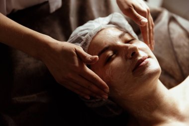 Bir kozmetik uzmanı genç bir kadının kafasında ve yüzünde güzellik prosedürü uygular..