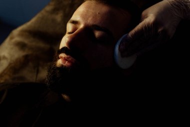 Güzellik uzmanında çeşitli güzellik tedavileri gören rahat genç bir adam sakallı bir adamın portresi güzellik iğneleri ve yüz maskeleri olan bir adam.