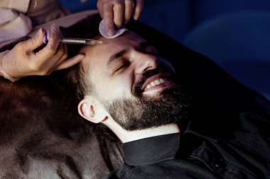 Güzellik uzmanında çeşitli güzellik tedavileri gören rahat genç bir adam sakallı bir adamın portresi güzellik iğneleri ve yüz maskeleri olan bir adam.