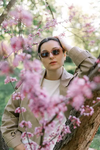 Bahar ceketi, pantolon ve güneş gözlüğü giymiş bir kadın, mülkünün parkında yürürken fotoğraf için poz veriyor.