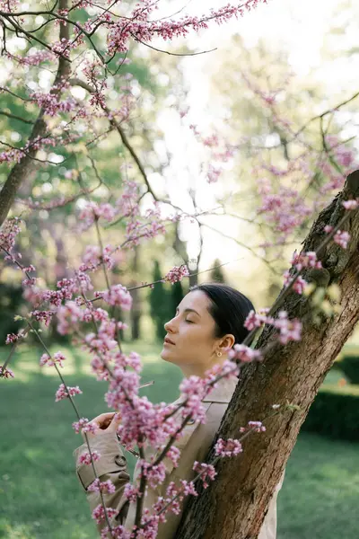 Bahar ceketi, pantolon ve güneş gözlüğü giymiş bir kadın, parkta çiçek açan bir kiraz ağacının arka planında zarif bir şekilde poz veriyor. 