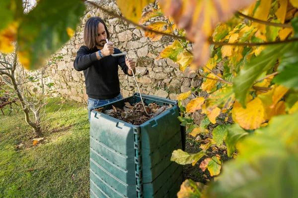 A man is mixing the organic waste with dry leaves in a outdoor compost bin placed in a garden to recycle home and garden wastes. Concept of recycling and sustainability