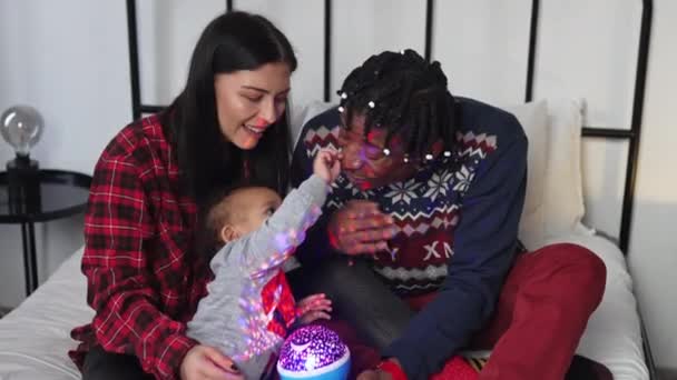 赤ん坊と一緒に若いカップルはクリスマスの前夜を楽しんでいます 赤ちゃんの夜のカラフルなランプで遊ぶ 高品質4K映像 — ストック動画