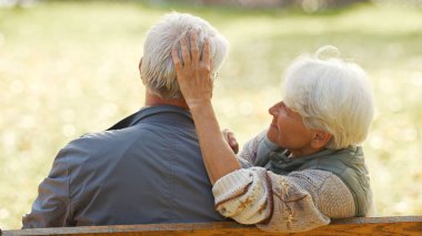 Back view of grey-haired senior woman sitting with her husband close together on a bench . High quality photo