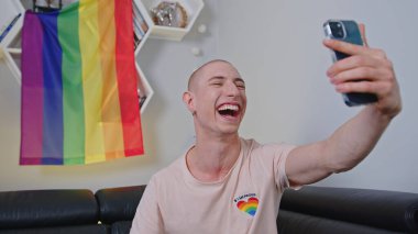 cheerful and satisfied boy laughing widely and taking a selfie with LGBT flag on the wall, medium closeup. High quality photo