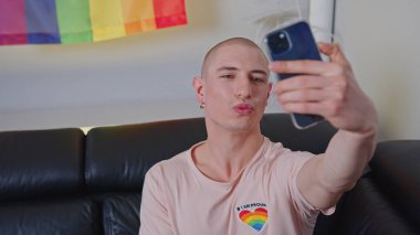 young Caucasian man in pink T-shirt taking a selfie in front of LGBT flag on the wall. High quality photo