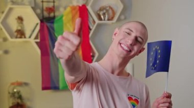 young Caucasian man with an earring holding the EU flag and showing thumb-up, LGBT flag in the background. High quality 4k footage