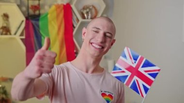 medium closeup shot of a young gay man with the UK flag showing thumb up, LGBT flag in the background. High quality 4k footage