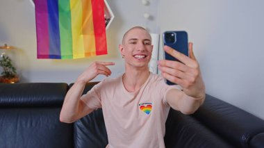 Queer young man taking selfie with LGBT flag in the background at home. High quality photo