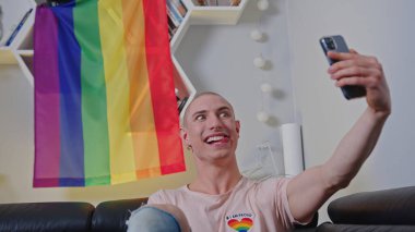 Smiling bald gay man taking selfie with LGBT flag in the background indoor. High quality photo