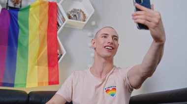 happy Caucasian boy with an earring and pink LGBT theme T-shirt doing a selfie shot, LGBT flag on the shelf, LGBT concept. High quality photo