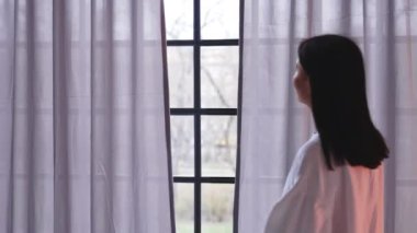 Back view of a woman opening window curtains and looking outside. High quality 4k footage