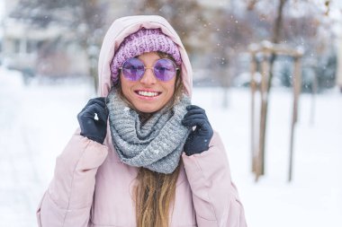 Smiling woman wearing sunglasses and winter clothes standing under the falling snow. High quality photo