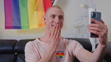 young man with an earing wearing pink LGBT theme T-shirt and looking surprised by the news in the phone, LGBT flag on the wall. High quality photo