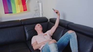 medium full shot of a young gay man on the sofa having a video call, LGBT flag on the wall. High quality 4k footage