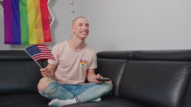 young homosexual man on the sofa enjoying the TV and holding the USA flag, LGBT flag behind her. High quality 4k footage