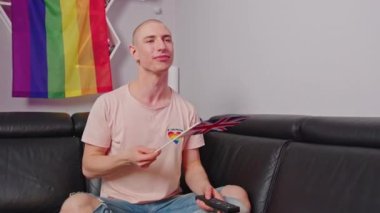 Caucasian gay man watching sitting on the sofa watching TV and waving the UK flag, LGBT flag on the background. High quality 4k footage