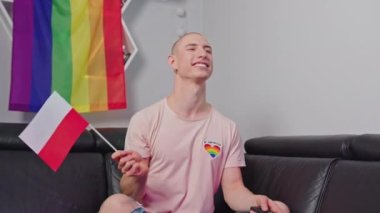 cheerful young man waving Poland flag and watching TV, LGBT flag on the shelves, LGBT concept medium closeup. High quality 4k footage