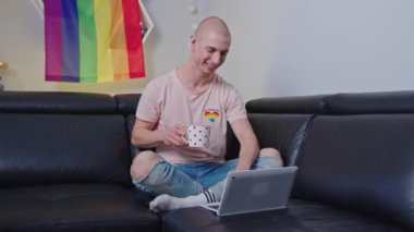 Caucasian boy sitting on the sofa drinking coffee and watching something funny on a laptop, LGBT. High quality 4k footage