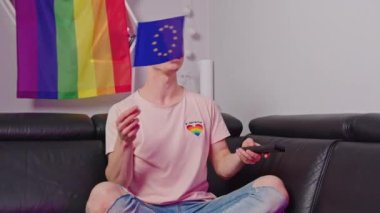 excited gay man in LGBT theme t-shirt waving EU flag while watching TV, LGBT flag. High quality 4k footage