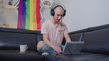 young bald gay man with headphones using a laptop and talking to somebody in the living room, LGBT flag on the wall. High quality 4k footage