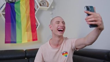 cheerful gay man recording a live video and greeting his viewers, LGBT flag hanging on the wall. High quality 4k footage