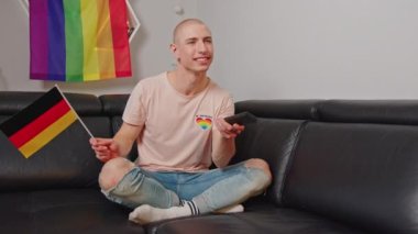 young man sitting on a sofa watching TV with a big interest and waving German flag, LGBT flag behind him. High quality 4k footage