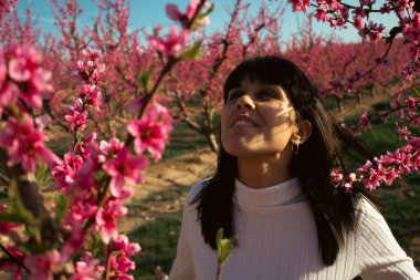 Baharda çiçek açan şeftali ağaçlarının arasında gülümseyen bir kadın.