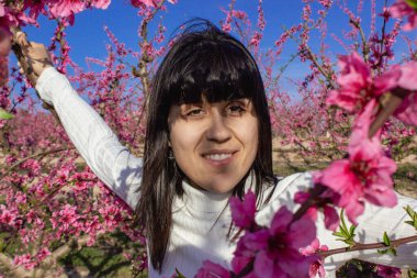 Baharda çiçek açan şeftali ağaçlarının arasında gülümseyen bir kadın.