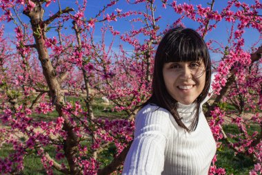 Baharda çiçek açan şeftali ağaçlarının arasında gülümseyen bir kadın.