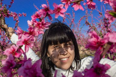 Baharda çiçek açan şeftali ağaçlarının arasında gülümseyen bir kadın.