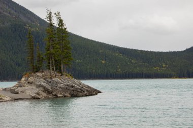 Küçük bir adada bulutlu bir gün ve Minnewanka Gölü boyunca büyüleyici bir dağ manzarası. Kanada Rocky Dağı, Banff Ulusal Parkı.
