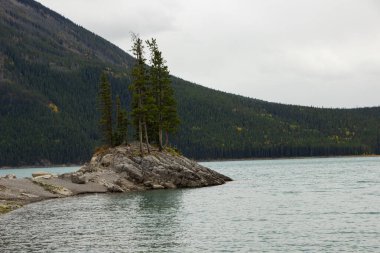 Küçük bir adada bulutlu bir gün ve Minnewanka Gölü boyunca büyüleyici bir dağ manzarası. Kanada Rocky Dağı, Banff Ulusal Parkı.