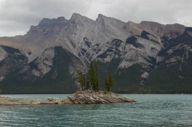 Küçük bir adada bulutlu bir gün ve Minnewanka Gölü boyunca büyüleyici bir dağ manzarası. Kanada Rocky Dağı, Banff Ulusal Parkı.