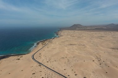 İspanya, Fuerteventura 'daki Corralejo Doğal Parkı kum tepelerini geçerken çekilmiş yüksek çözünürlüklü hava fotoğrafı. Görüntü, altın kum tepeciklerinden oluşan engin bir çöl manzarasını kesen düz bir asfalt yolu gösteriyor.