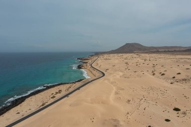 İspanya, Fuerteventura 'daki Corralejo Doğal Parkı kum tepelerini geçerken çekilmiş yüksek çözünürlüklü hava fotoğrafı. Görüntü, altın kum tepeciklerinden oluşan engin bir çöl manzarasını kesen düz bir asfalt yolu gösteriyor.